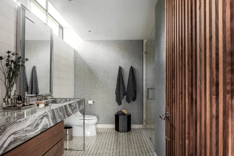 A long pale veined counter over wood drawers faces a vertically slatted wood wall beside light gray walls, a white toilet and small floor tiles