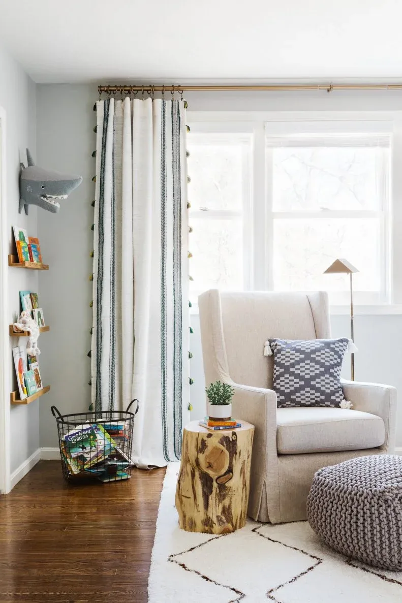 Pale adult armchair beside wood-toned cylindrical table, textured pouf, book basket and finer patterned curtains.