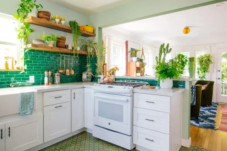 White kitchen with a green tiled backsplash, wood shelves, hanging utensils and leafy plants.