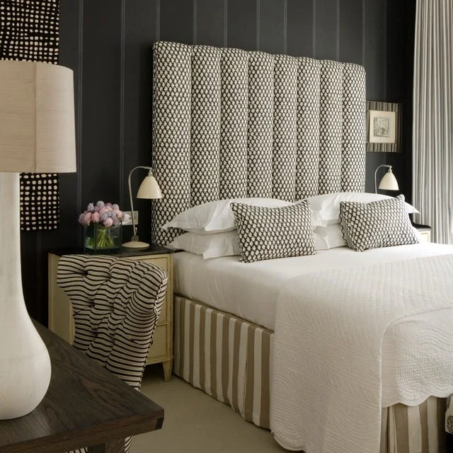 A tall black-and-white patterned headboard stands against a dark paneled wall beside small pale lamps, striped bed upholstery and a curved-pattern chair