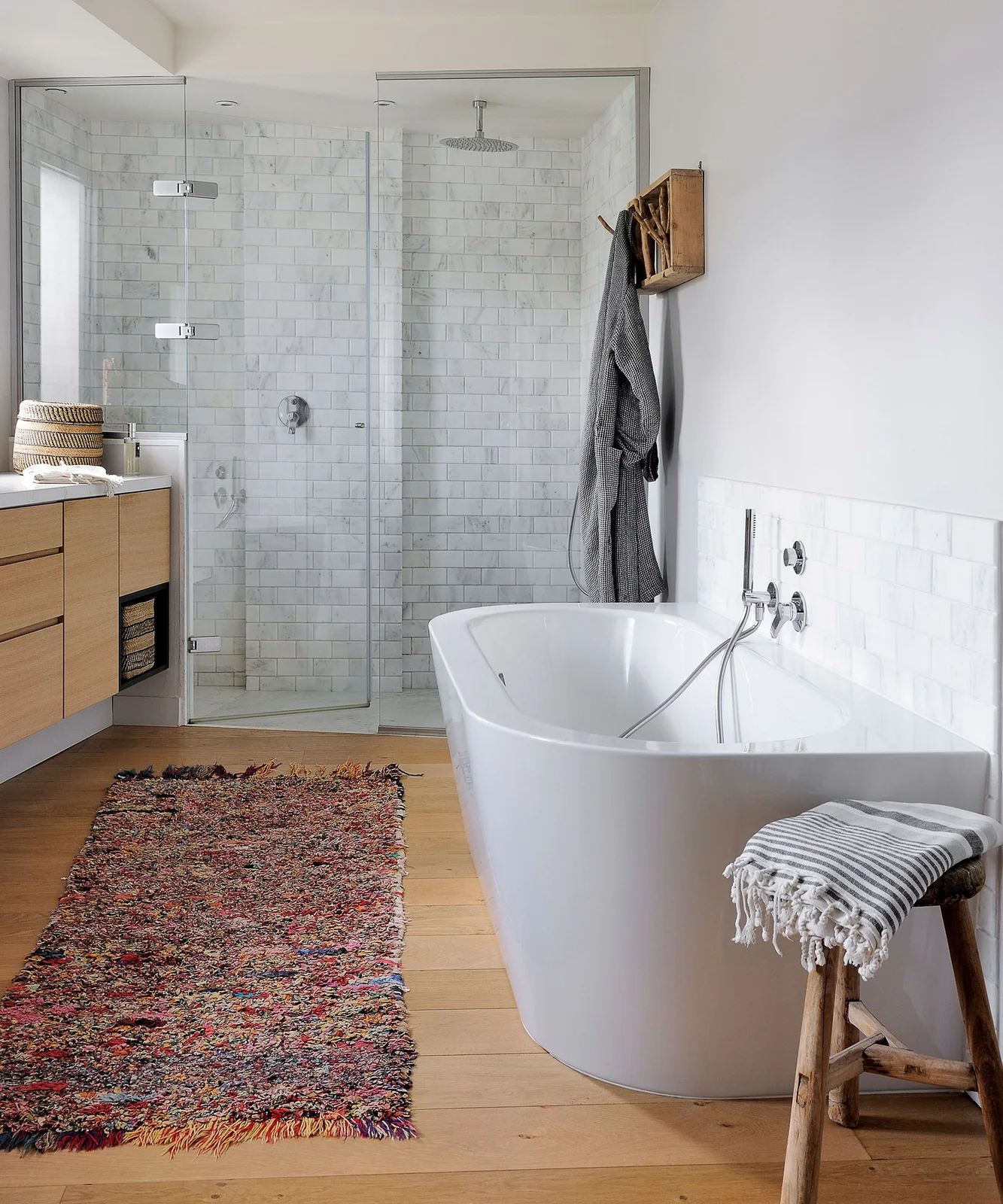 A white freestanding bath stands beside a clear shower enclosure and wood-front vanity over warm flooring with a patterned rug and small wood stool