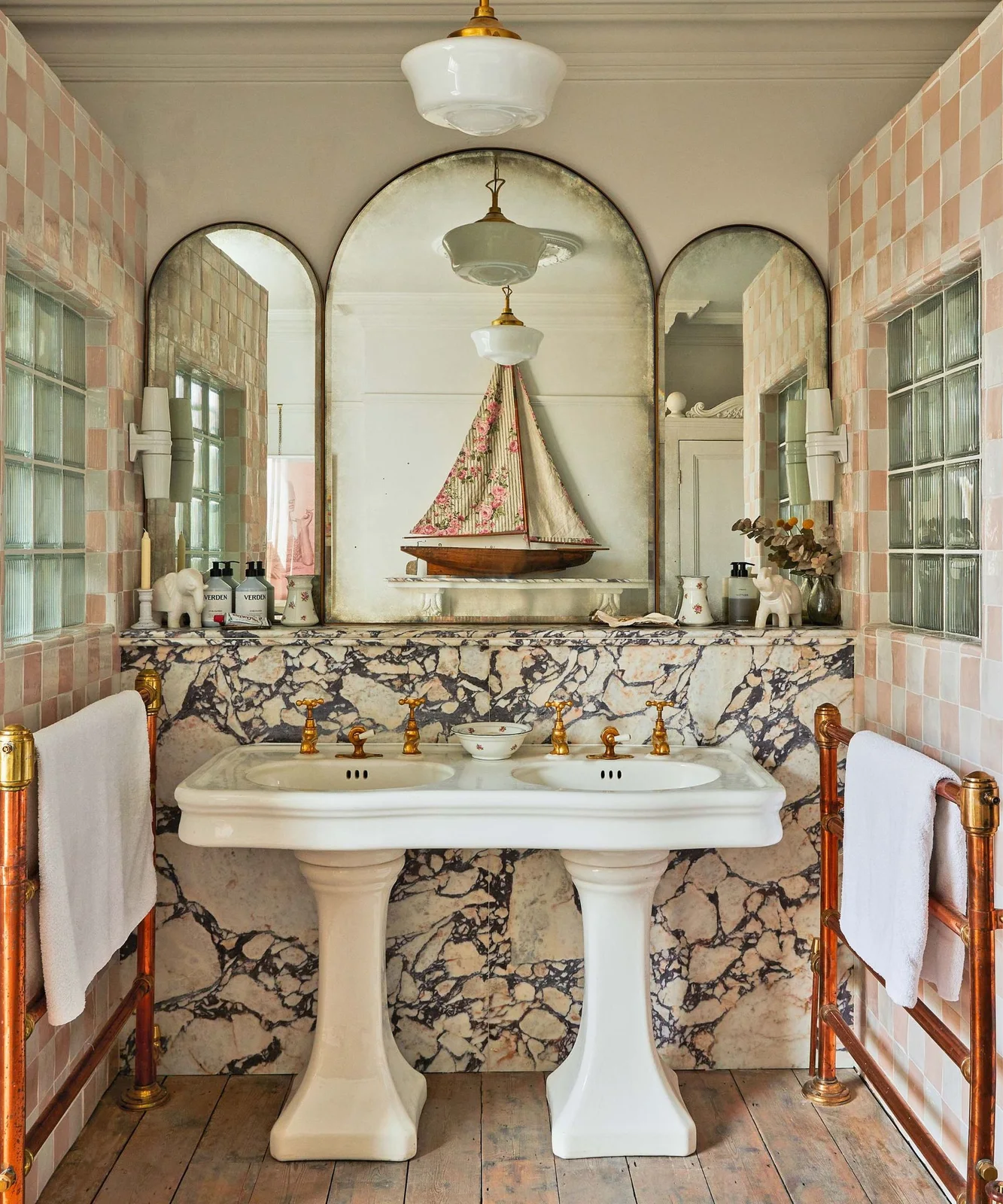 A double pedestal basin sits beneath three arched mirror sections against veined lower walls and pink checked tiles