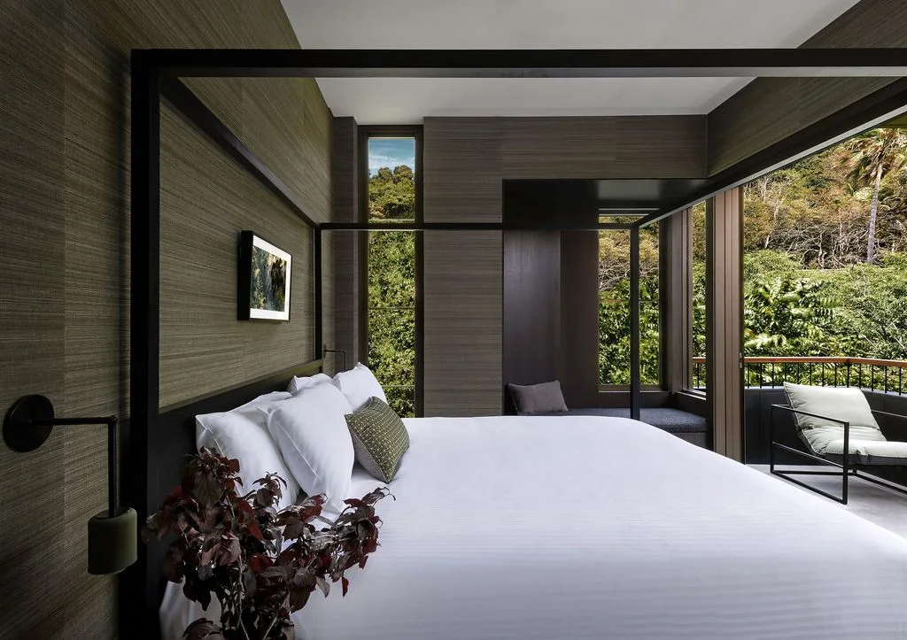 A dark open four-poster bed with white bedding stands against muted textured walls beside broad dark-framed glazing and a narrow side surface