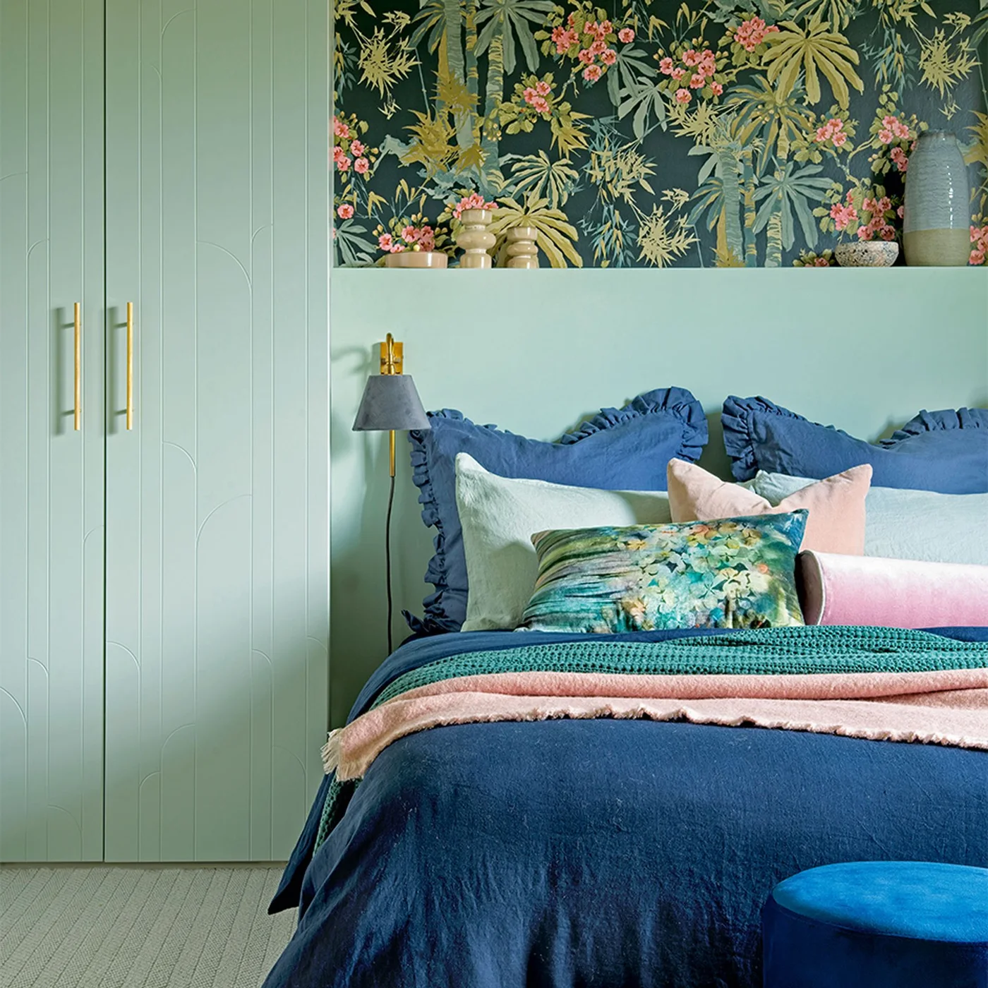 Pale green cabinets and a broad headboard sit below a darker botanical wall panel beside blue bedding and smaller pink accents