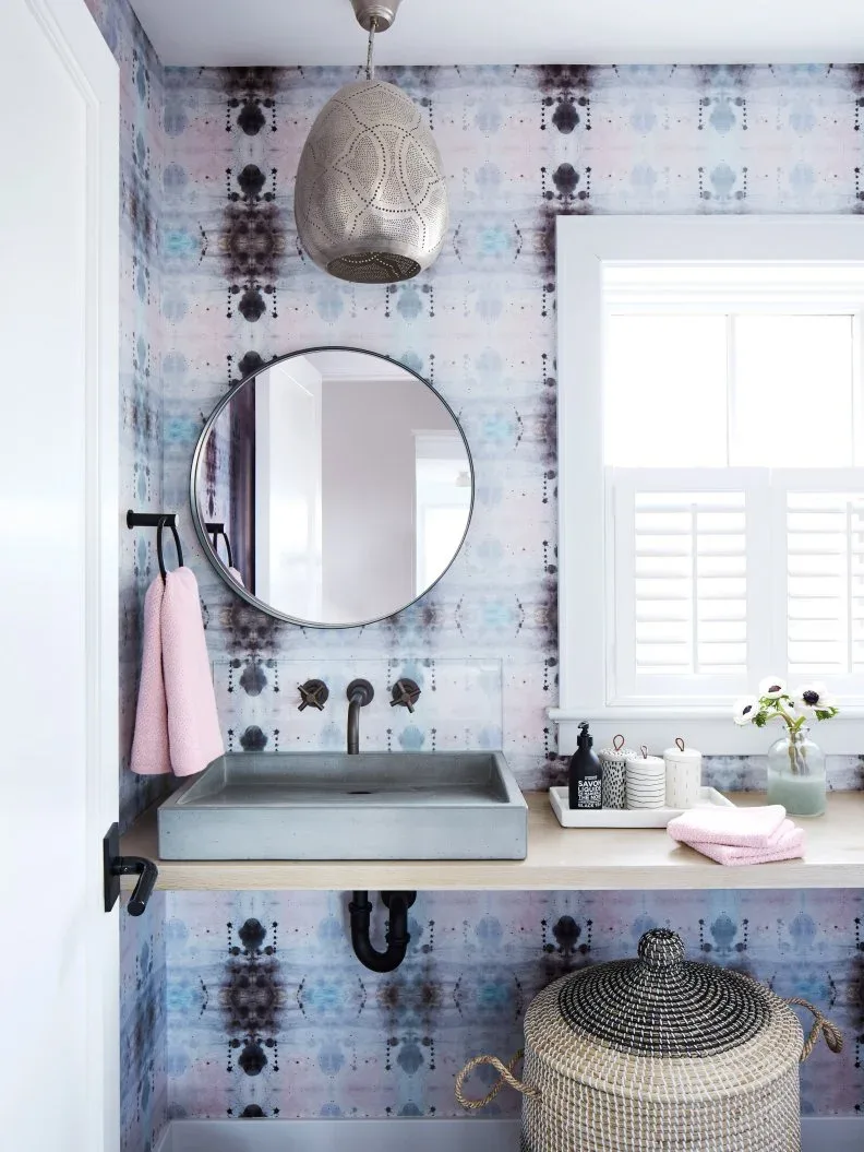 Powder room with pale abstract patterned wallpaper, round dark mirror, gray rectangular basin, open pale counter, textured pendant and large lidded woven basket.