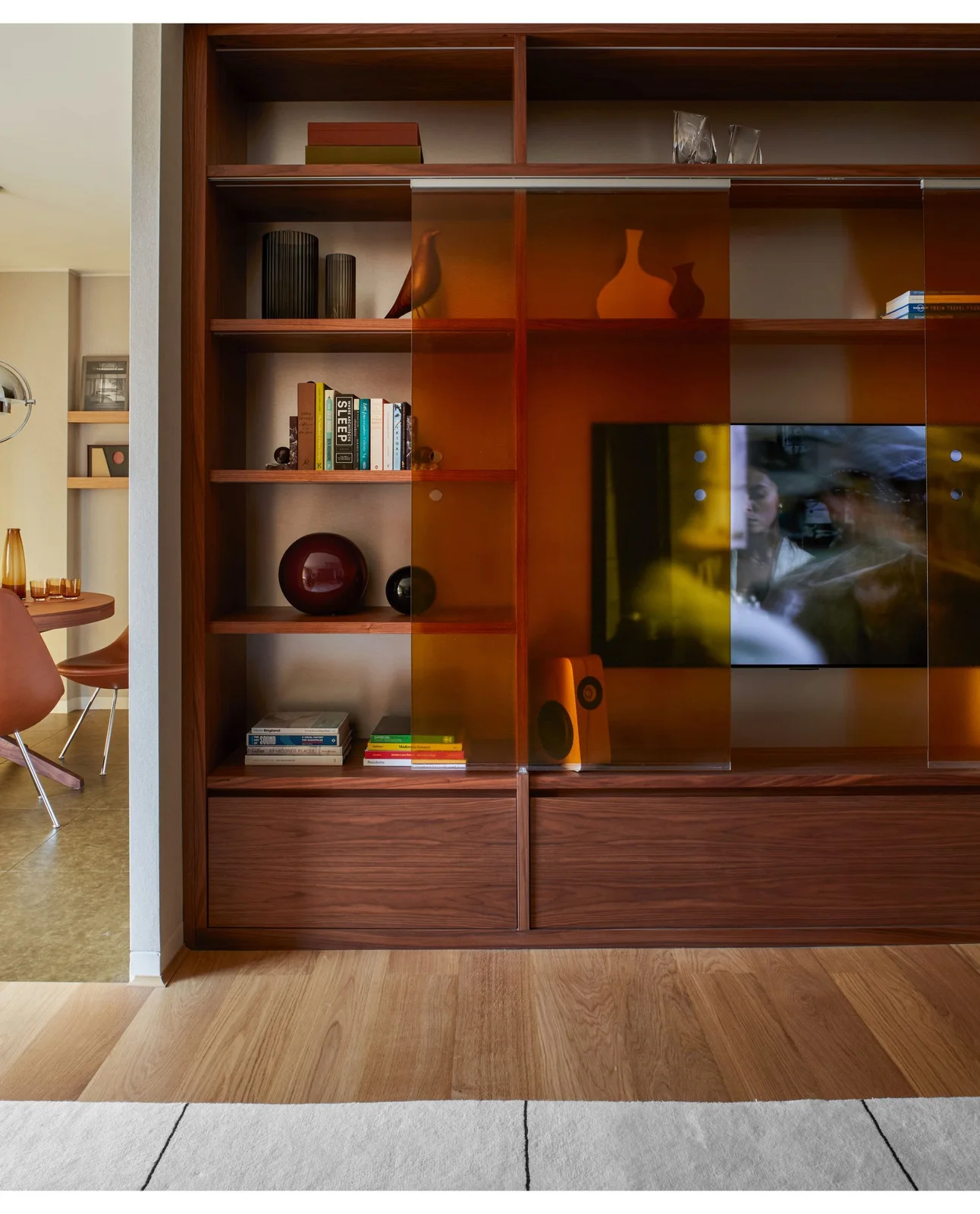 Amber sliding-glass panels cross wooden media shelves containing a television, books and decorative vessels above lower drawers