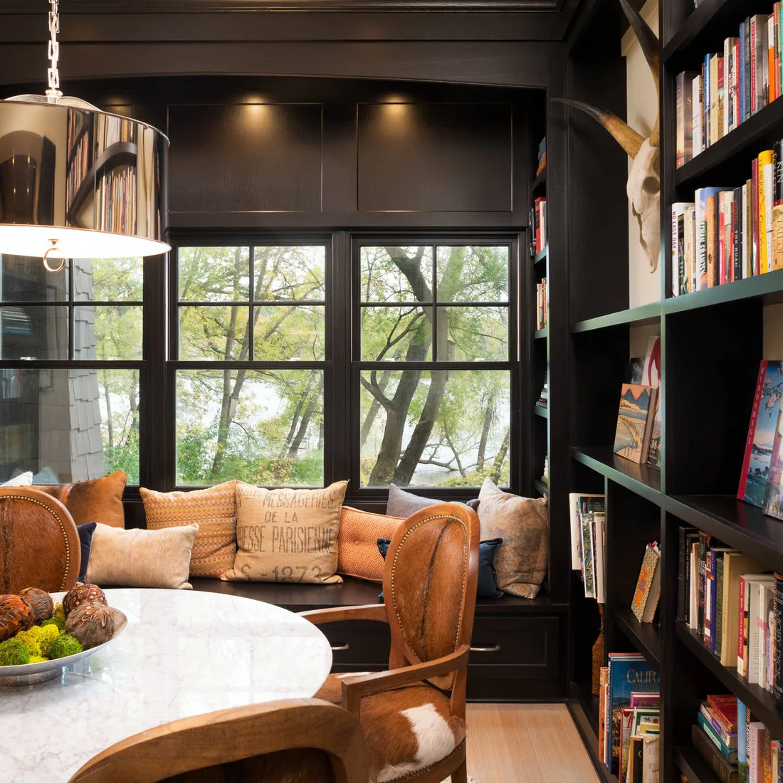 Dark bookshelves beside a window dining bench with warm-toned chairs and a pale round tabletop