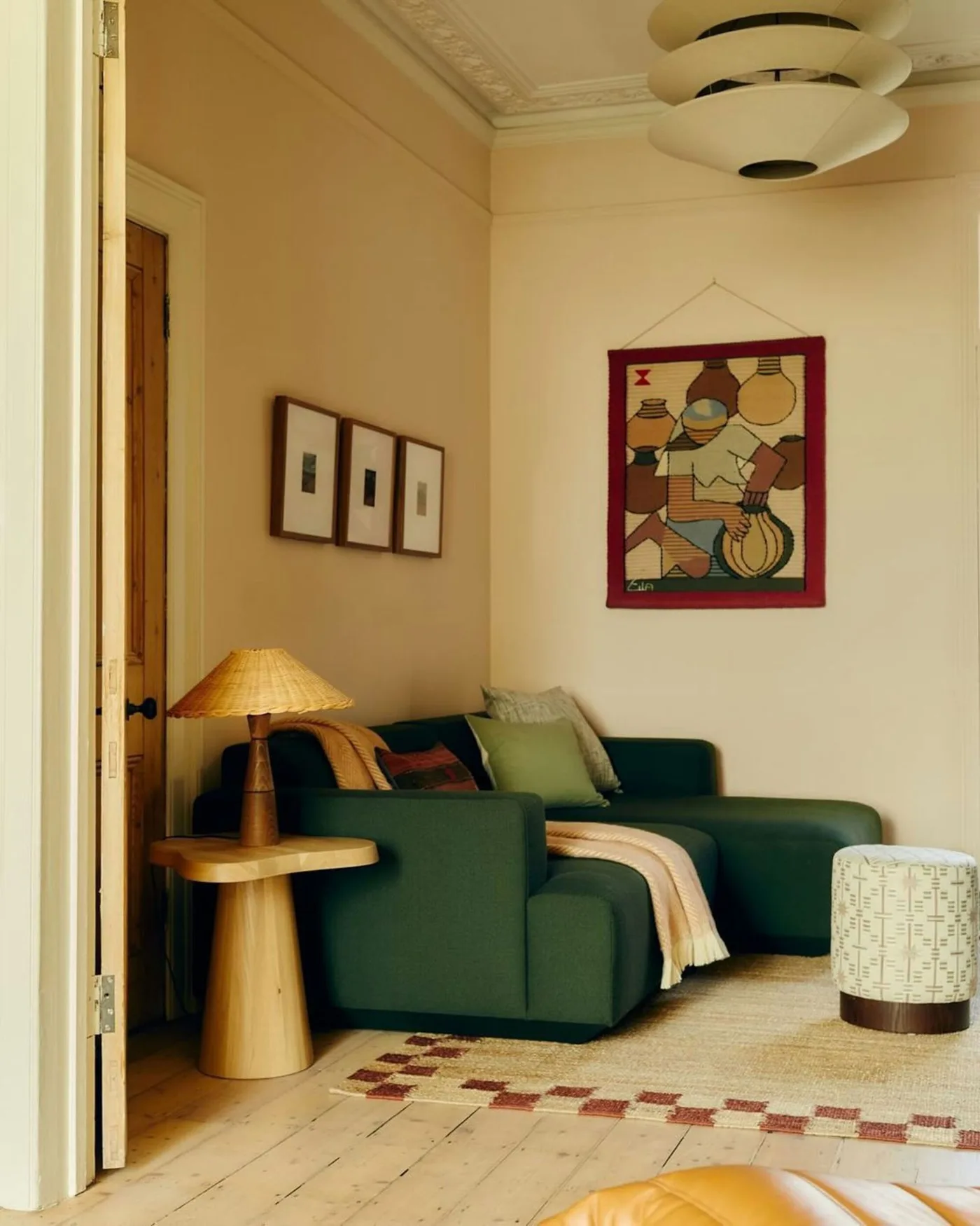 A deep green corner sofa sits beneath framed artwork beside a wooden side table and warm-toned lamp