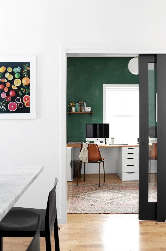 A wooden desk with pale drawer units and a brown chair sits against a deep green wall beneath a narrow shelf