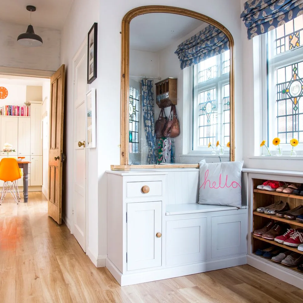 White entryway bench and cupboard beneath an arched gold-colored mirror beside open shoe cubbies and patterned window blinds.