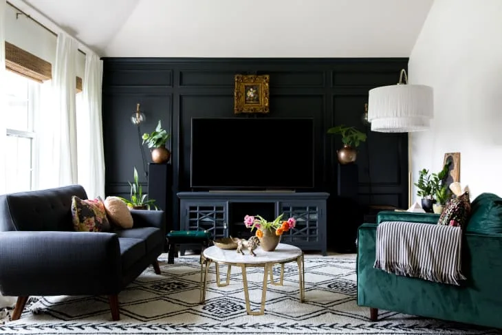A television and dark cabinet sit against a black paneled wall beside dark sofas, a round pale table and patterned rug