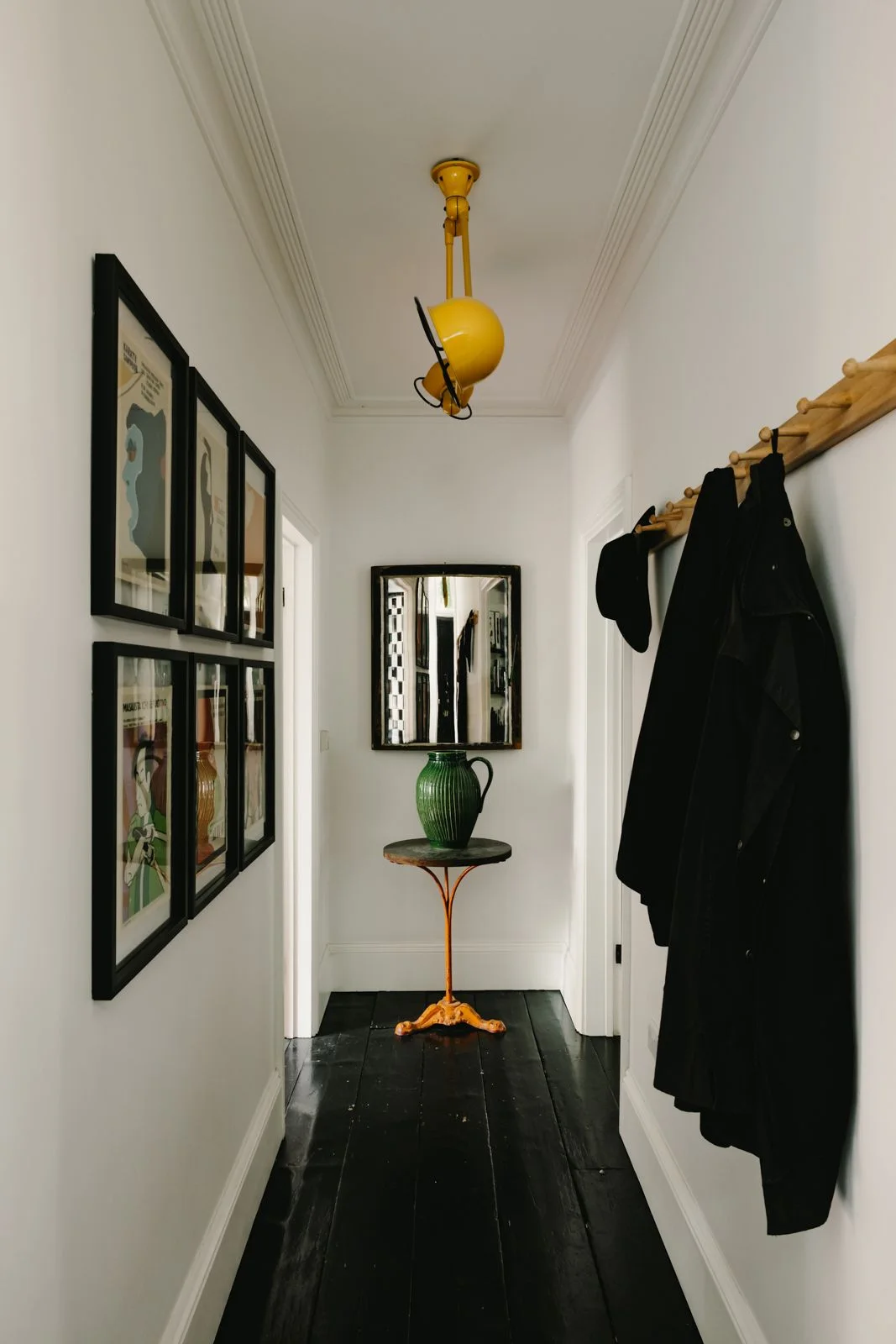 A narrow pale hallway has dark floorboards, black-framed artwork, a wooden coat rail with dark coats and a small round table beneath a yellow light