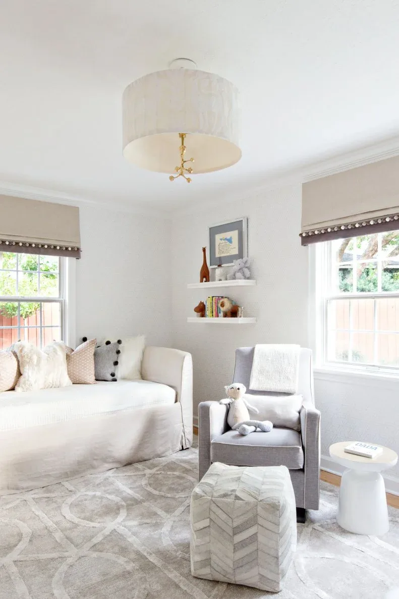 Light nursery seating area with pale daybed, gray chair, patterned ottoman, small table and window shades