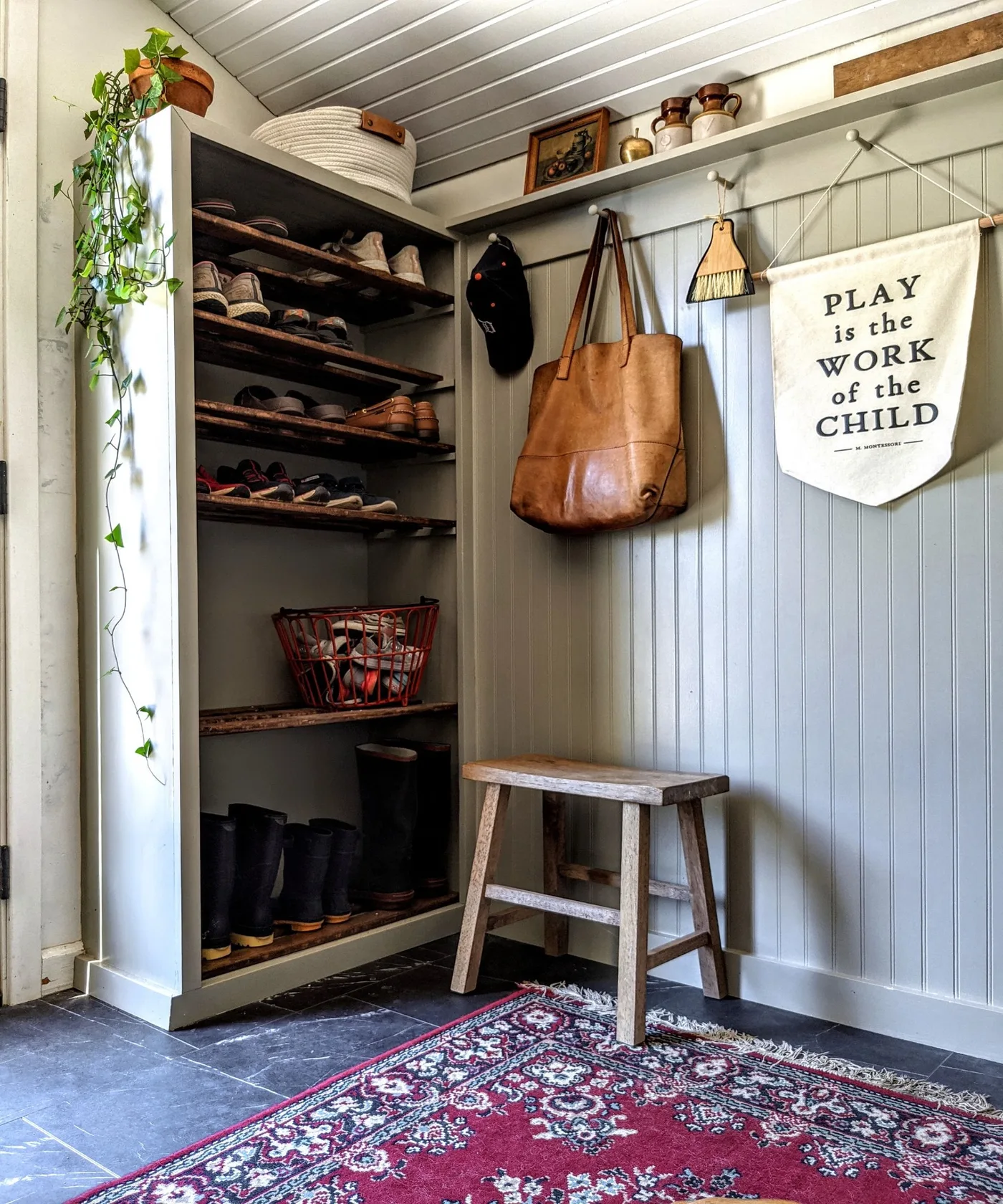 Entry corner with pale paneling, tall open shoe shelves, small wood stool, coat hooks, upper display shelf and reddish patterned rug