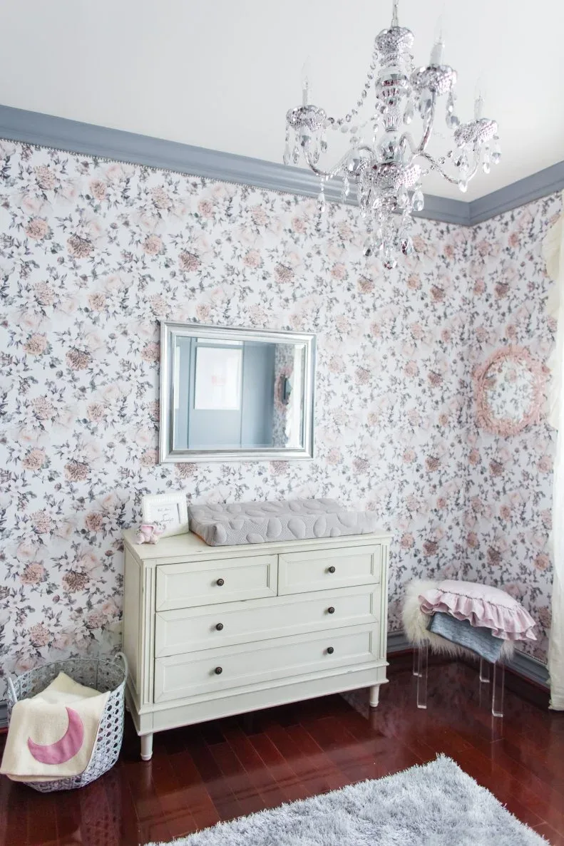 Pale dresser against small floral wallpaper with gray trim, rectangular mirror, decorative chandelier and smaller upholstered seat.