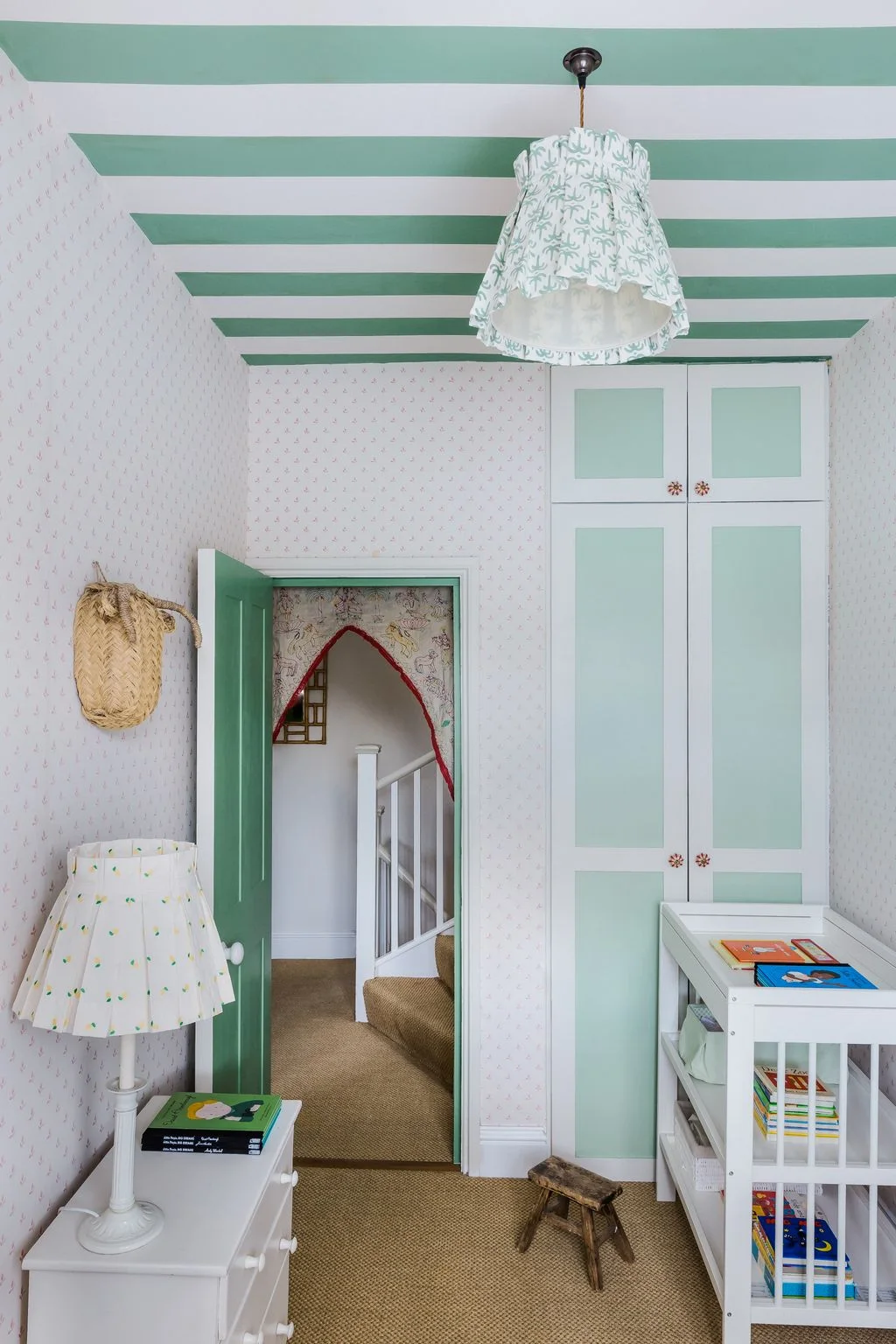 Green-and-white ceiling stripes sit above fine nursery wallpaper beside green paneled storage, a white crib and patterned lampshades