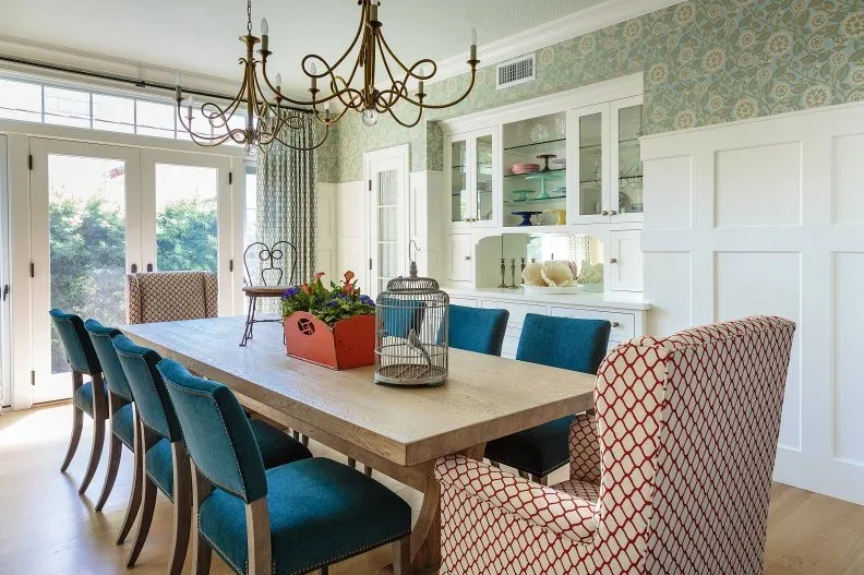 White fitted dining-room cabinets with glazed upper doors and a recessed serving area, beside patterned wallpaper, a wooden table, blue chairs, and red-patterned end chairs.