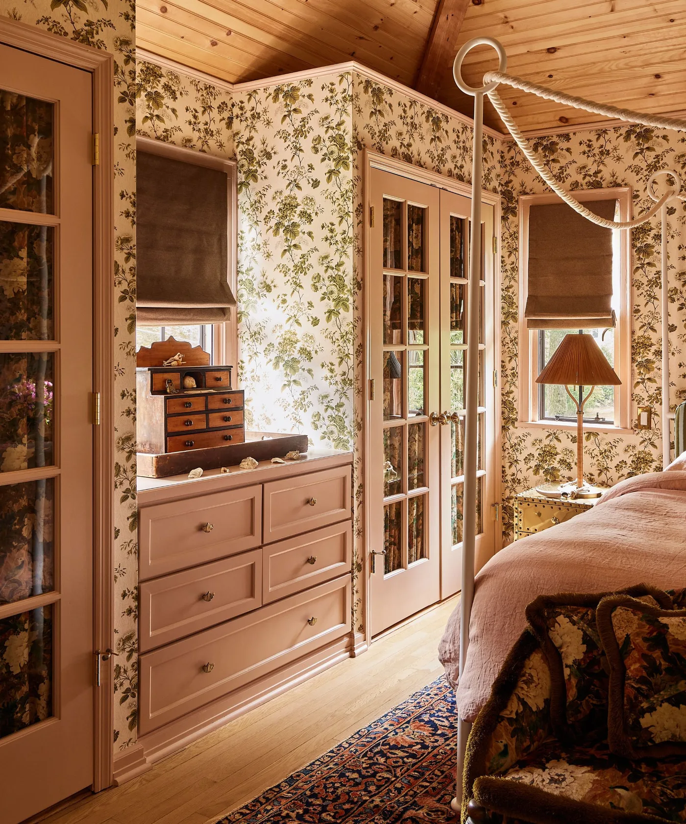 Green floral wallpaper around pink bedroom drawers, glass-fronted cupboards and a window beside a canopy bed.