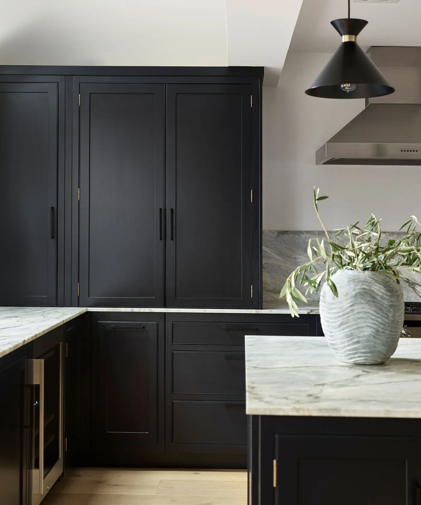 Kitchen with tall black paneled cupboards, dark lower drawers, pale veined island and counters, black pendant, metal hood, and warm wood floor.