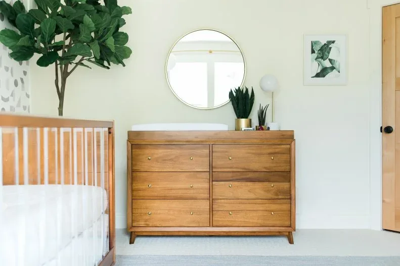 A wooden crib and dresser sit against pale green walls beside a round mirror and a large leafy plant