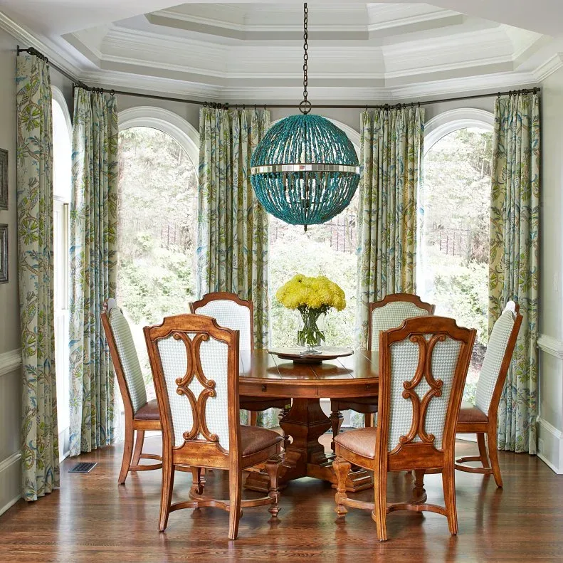 Round wooden dining table with decorative chair backs, floral curtains and a blue-green spherical light in a window bay.