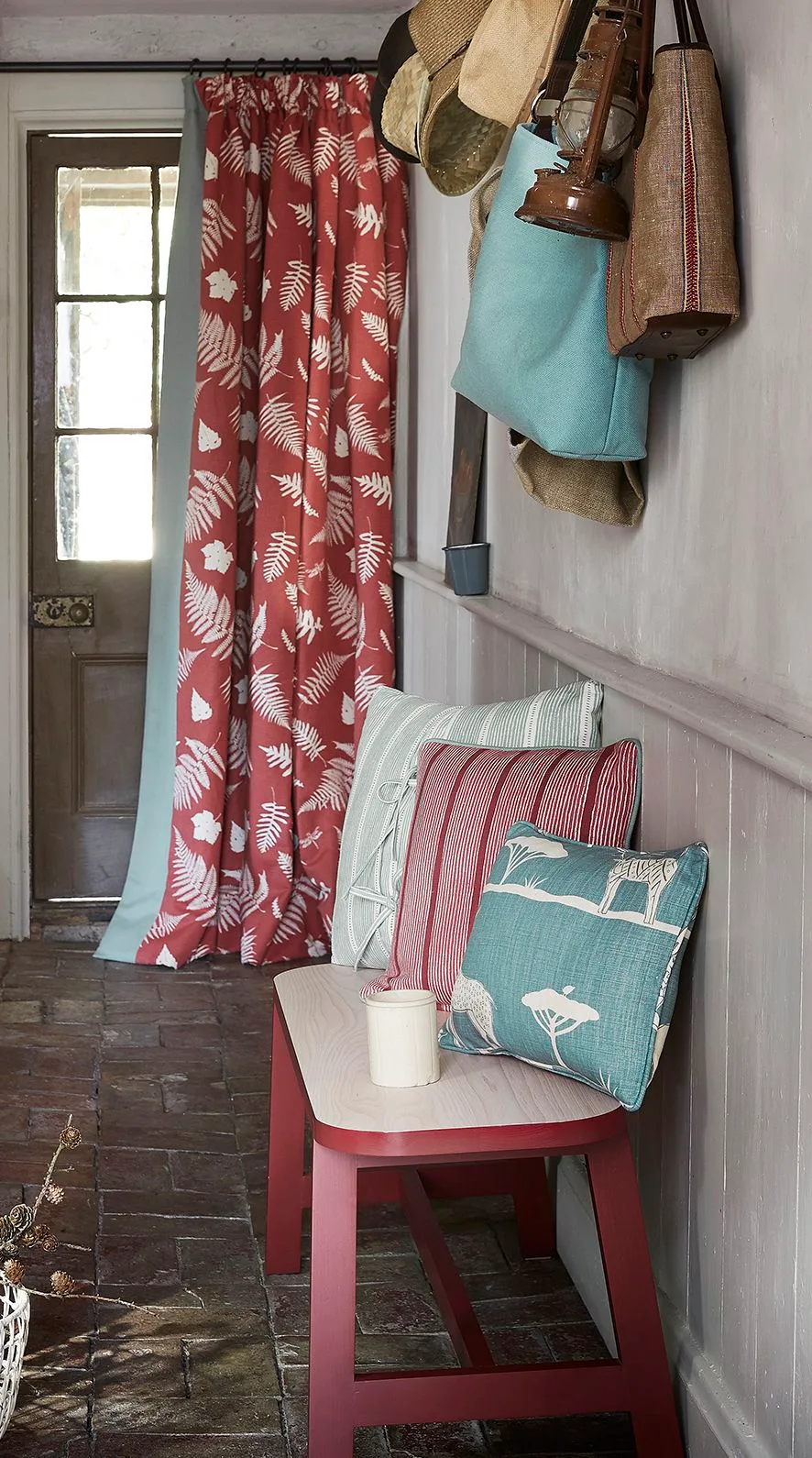 Country entryway with a red leafy door curtain, small bench with red legs, patterned cushions and hats and bags hanging above pale wall paneling.