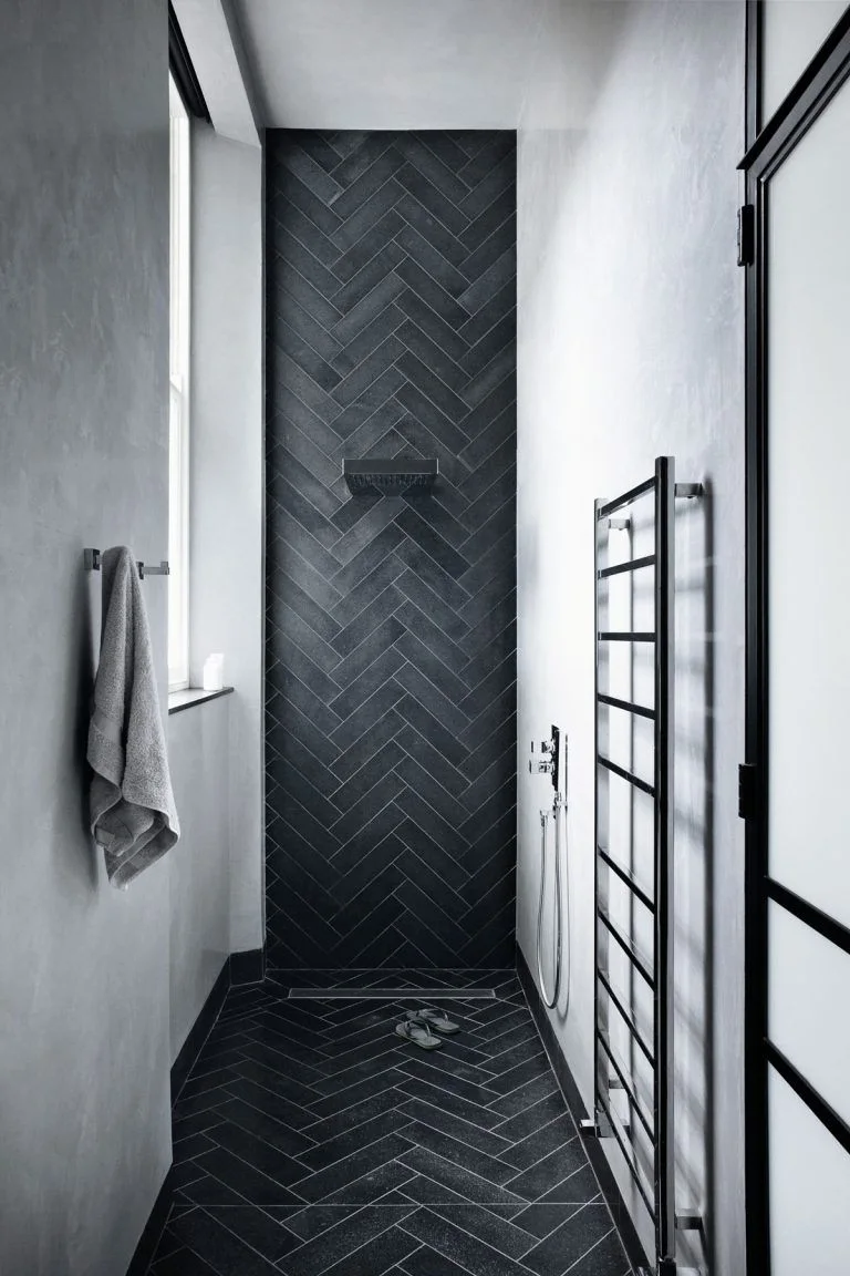 A narrow shower has dark herringbone tile at the far wall and floor beside pale walls, black fittings and a black ladder-style towel rail