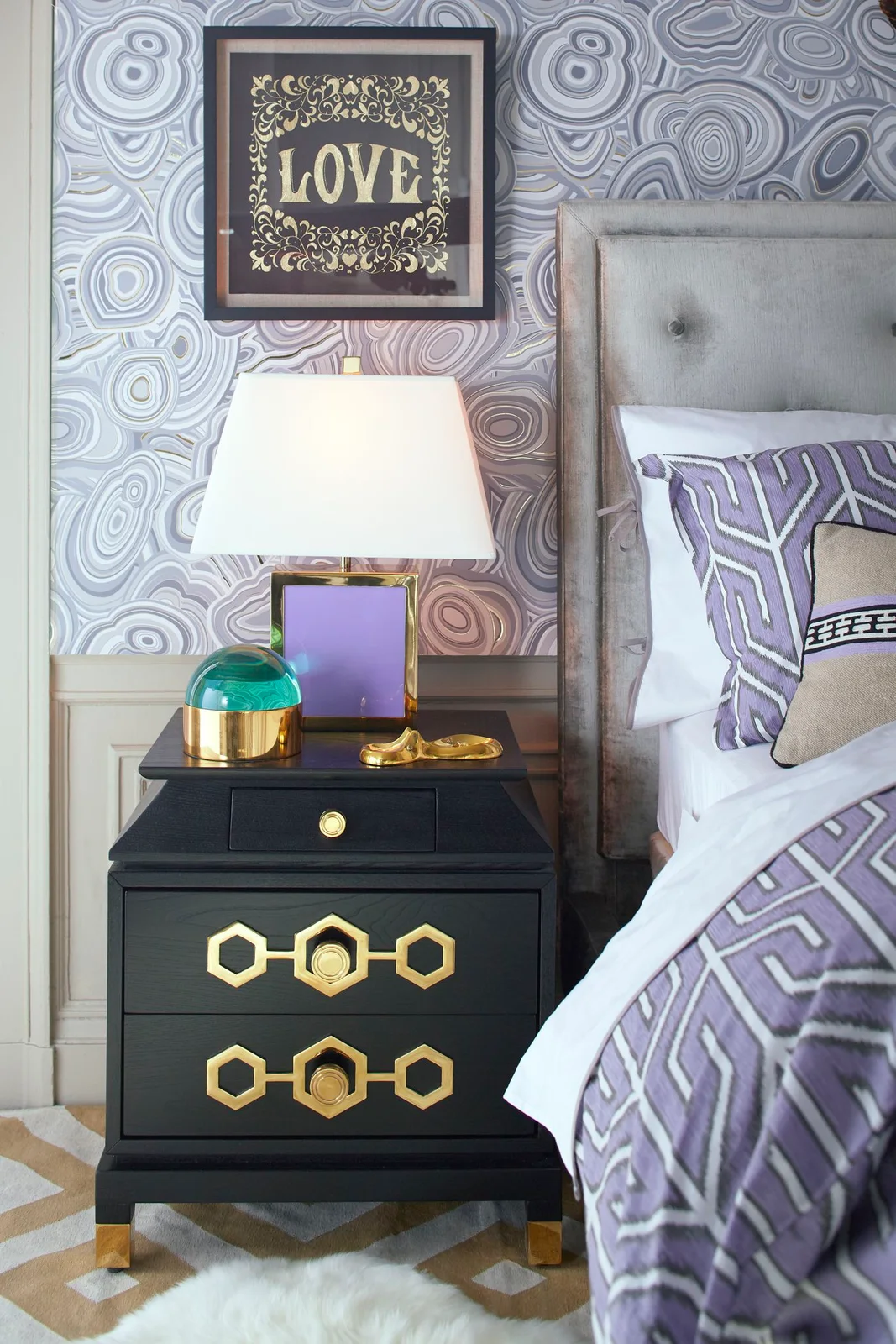 Lavender-gray flowing wall pattern surrounds a tufted headboard above purple geometric bedding beside a dark cabinet with gold-toned handles