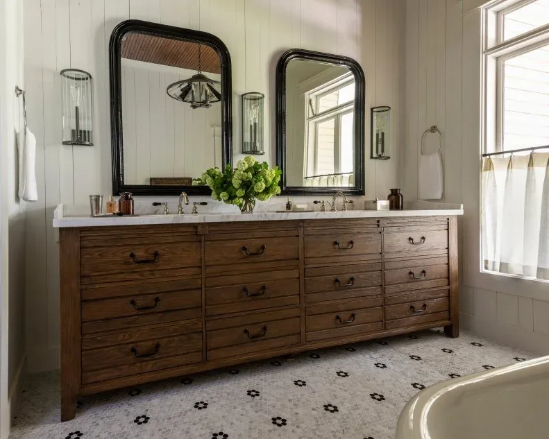 A wood double vanity with many drawers sits beneath two large dark-framed mirrors and glass wall lights beside pale vertical wall boards and small patterned floor tiles