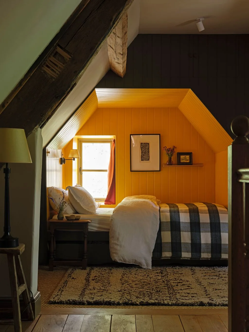 Attic bedroom with a yellow paneled bed recess, dark surrounding wall, exposed beams, checked blanket and small window