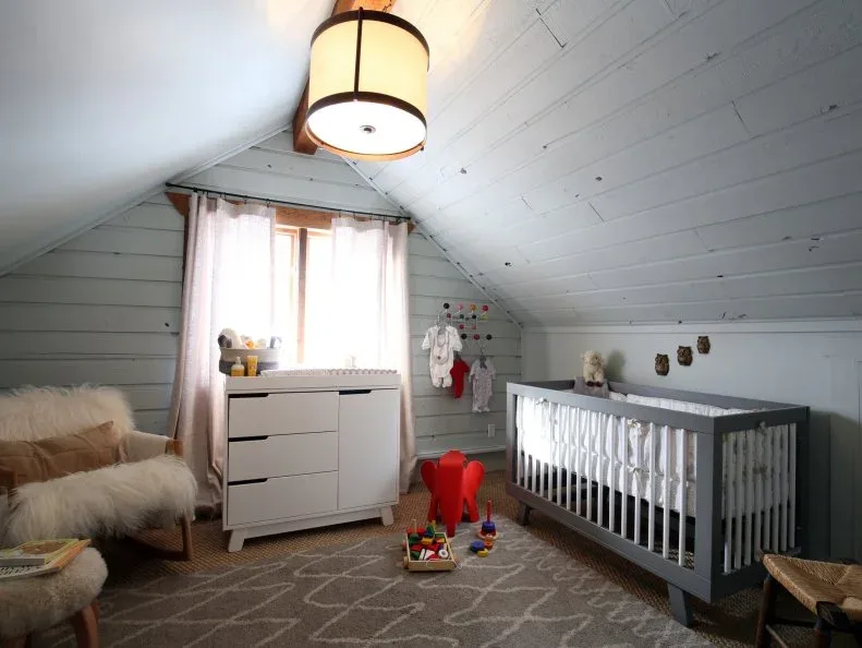 Gray paneled nursery with sloping ceiling, warm window trim, pale storage, simple crib and patterned rug.