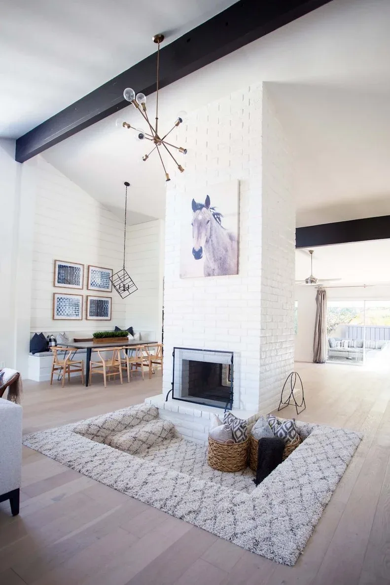 Tall white brick chimney between a dining area and pale sitting area beneath dark ceiling beams.