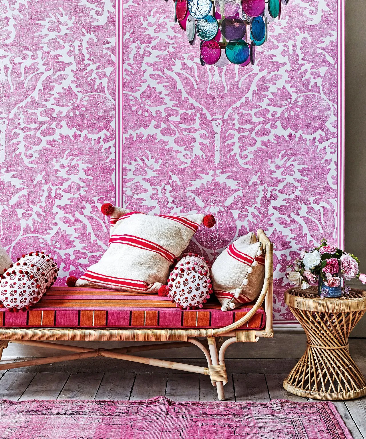 A woven daybed sits against pink patterned wallpaper beside a small natural table.