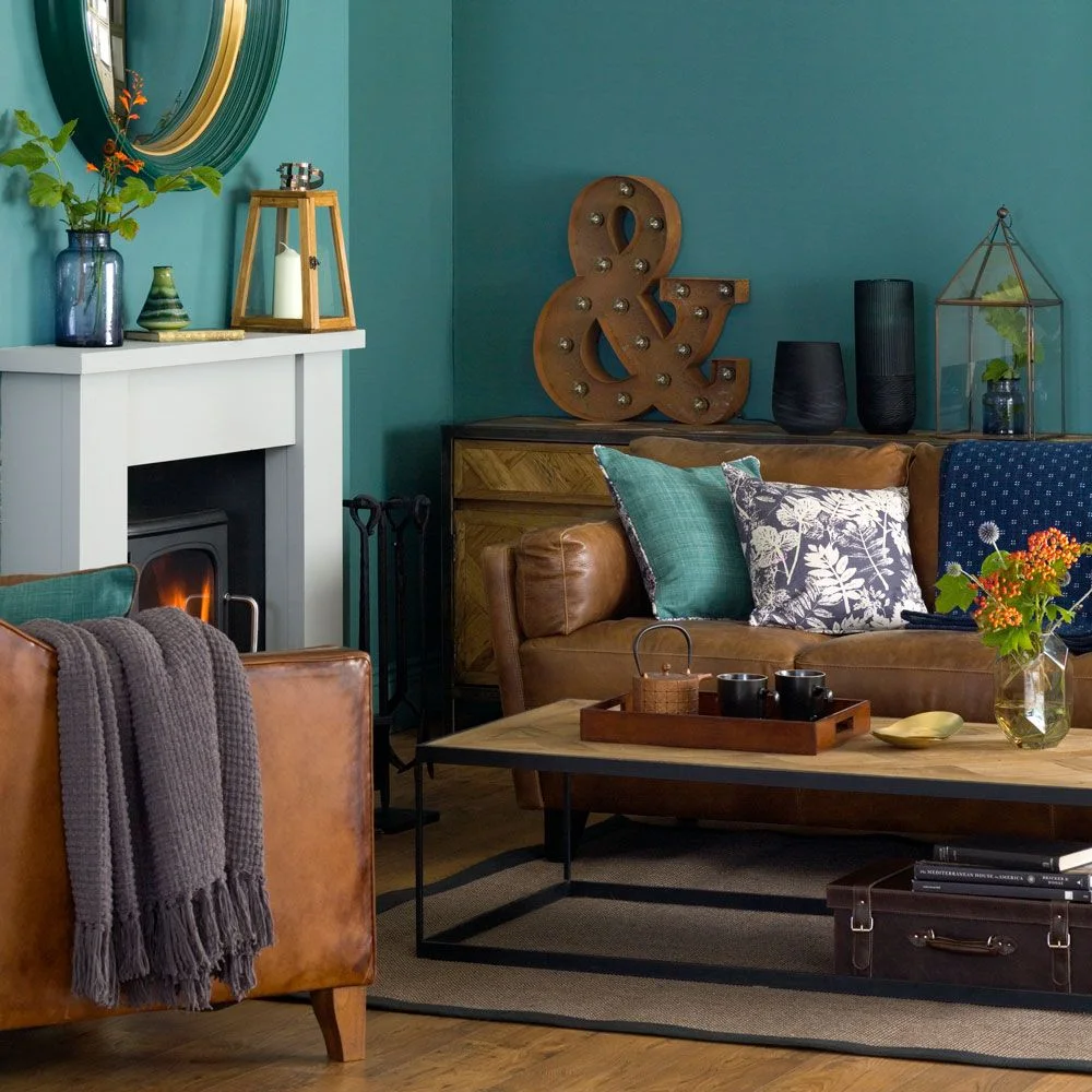 Brown seating and a wooden coffee table sit beside teal walls, a pale fireplace surround and patterned cushions