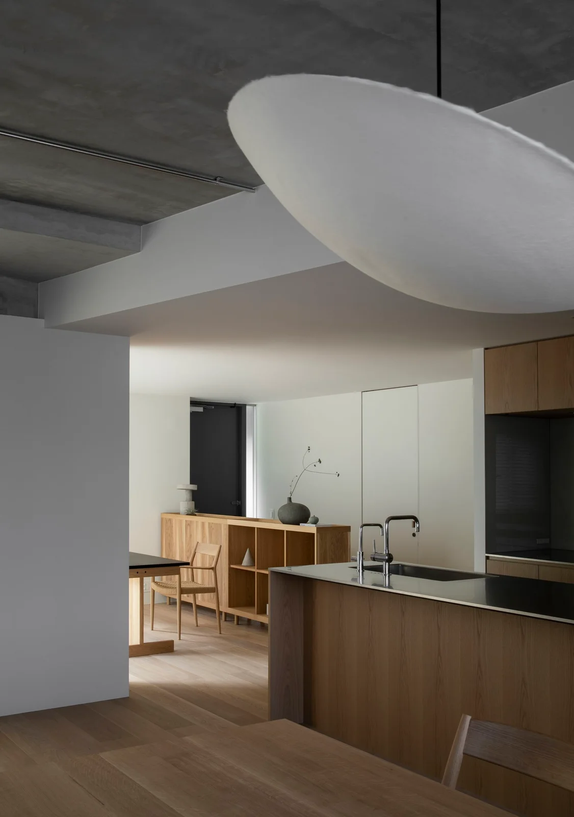 A wooden kitchen island with a dark counter stands beside a wooden dining chair and open shelf unit beneath a large pale pendant