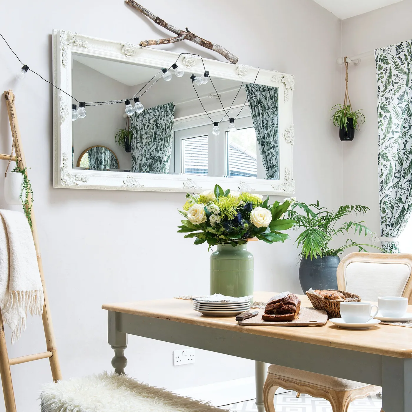 Ornate white rectangular mirror above a pale wood dining table with festoon lights and botanical curtains