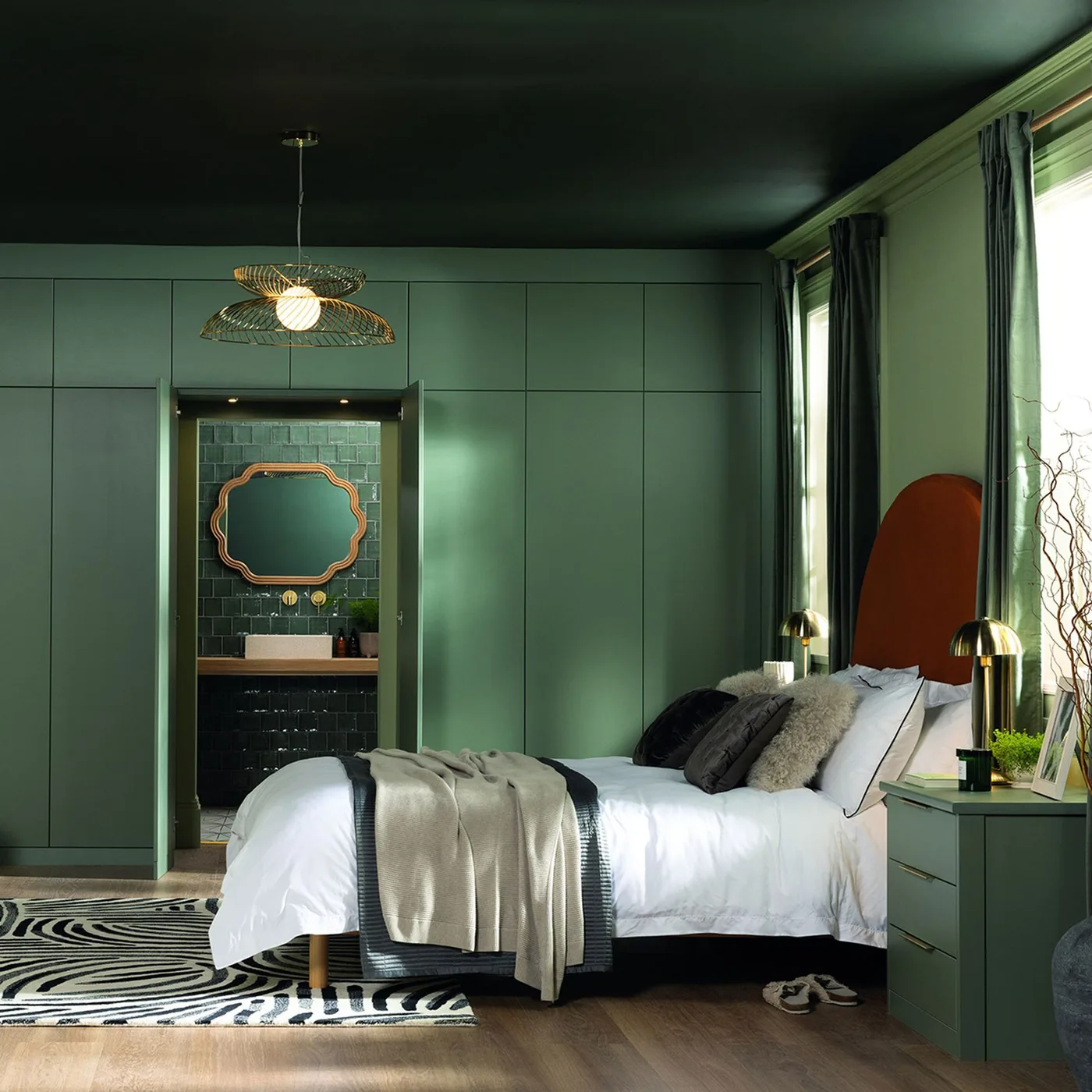 Bedroom with deep green ceiling, green fitted wardrobes, rust-colored curved headboard, gray bedding, dark curtains, and a doorway to a tiled bathroom.