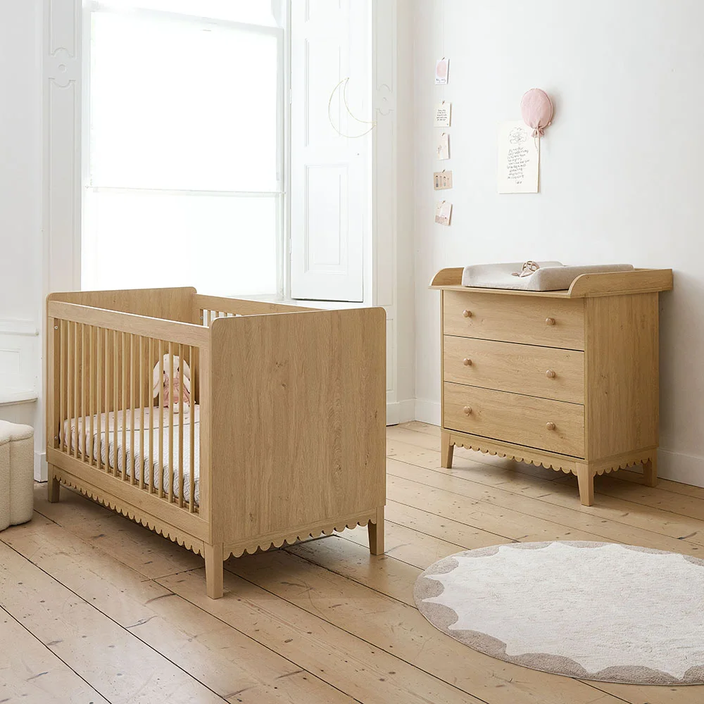 A pale wooden crib and matching drawer chest sit beneath a window in a white nursery with a warm wooden floor