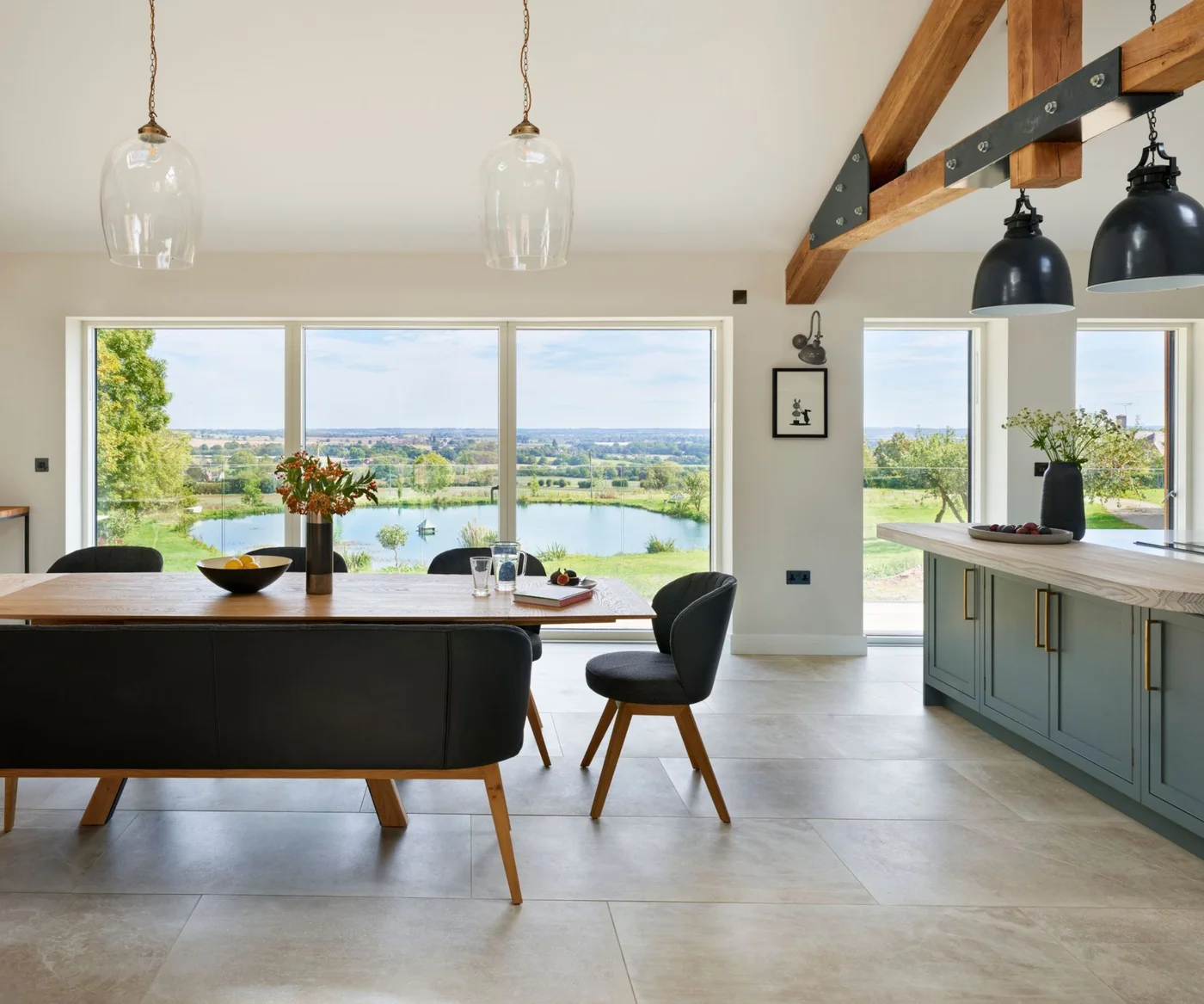 A wooden dining table and dark upholstered bench sit beside broad glazing, a green kitchen island and exposed wooden beams
