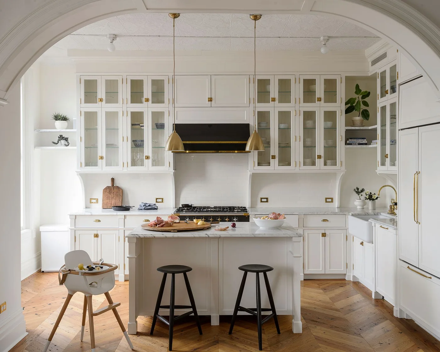 Pale framed kitchen cupboards with glass upper doors surround a small paneled island beneath slim warm-toned hanging lights and an arched room opening