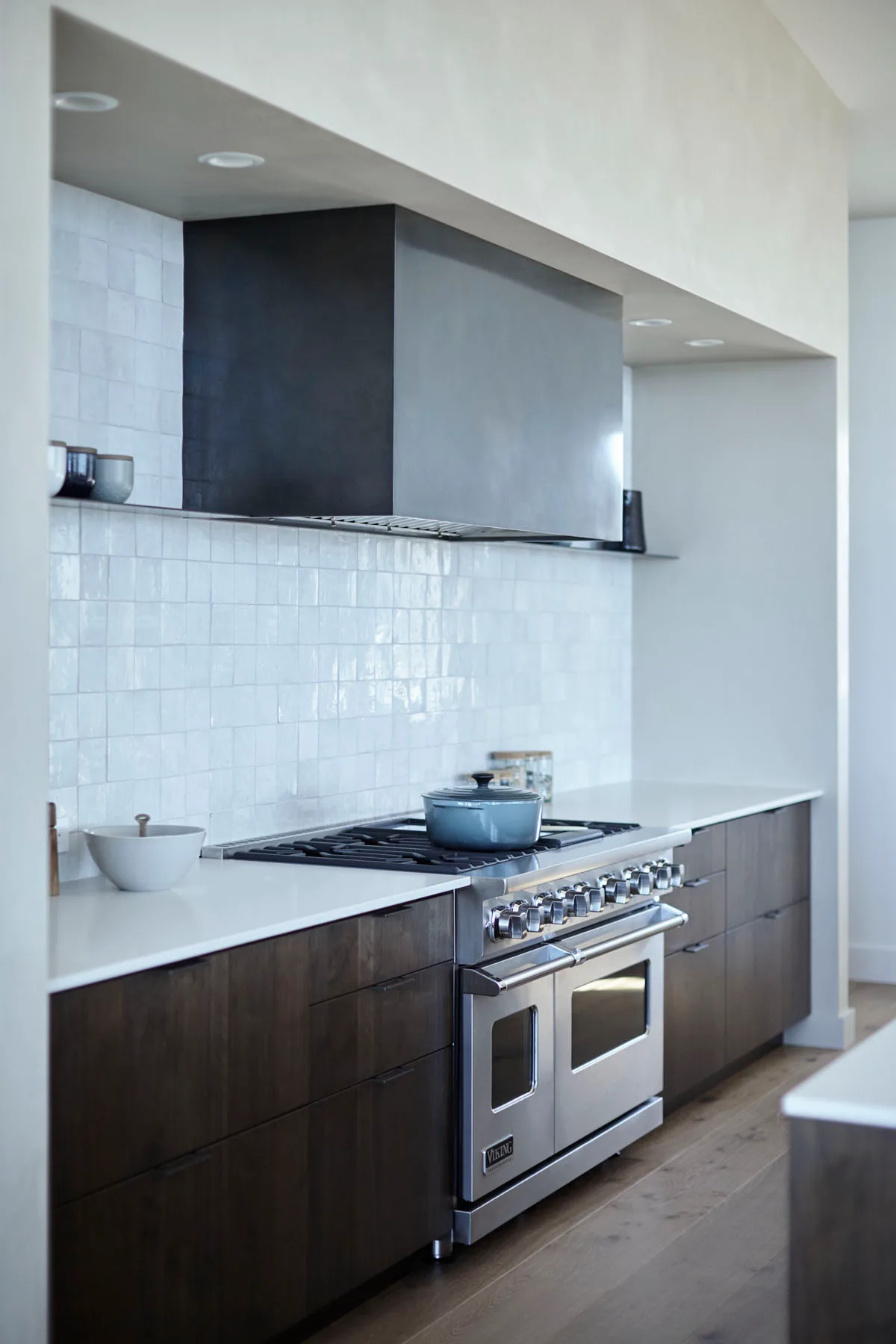 Kitchen with dark brown lower drawers, white counter and glossy square backsplash tiles, metal range, broad dark hood, and pale walls.