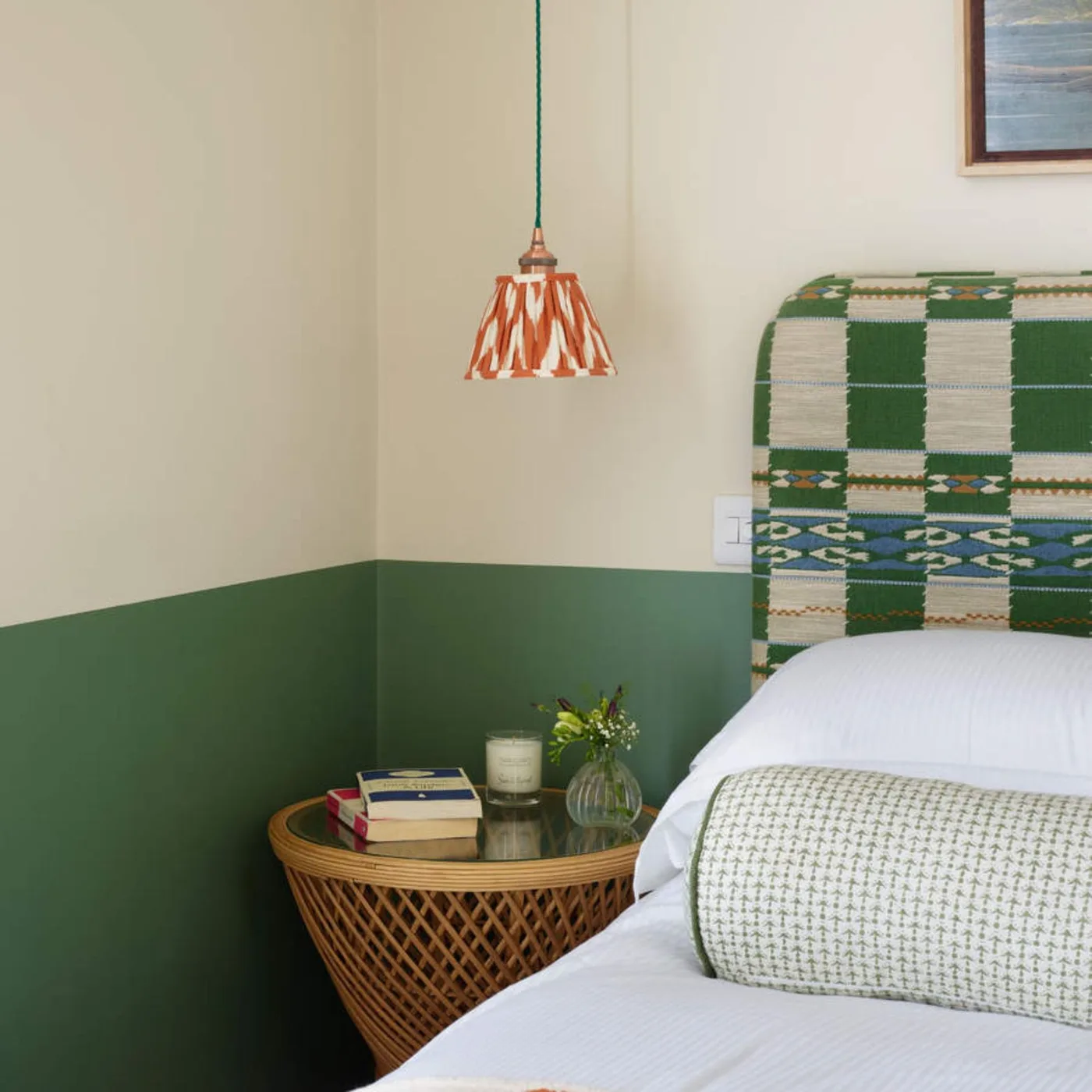Bedroom with a deep green lower wall, pale upper wall, patterned upholstered headboard, orange printed pendant, and round woven bedside table.