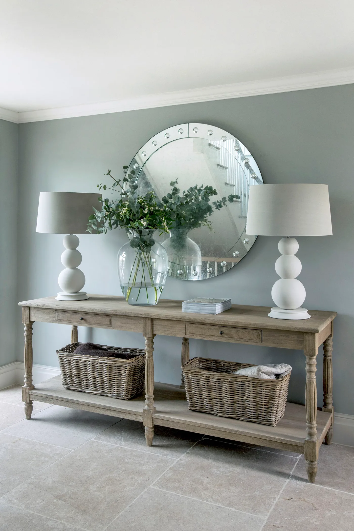 Pale green entryway with a round mirror, paired white lamps and a wooden console with two baskets below.