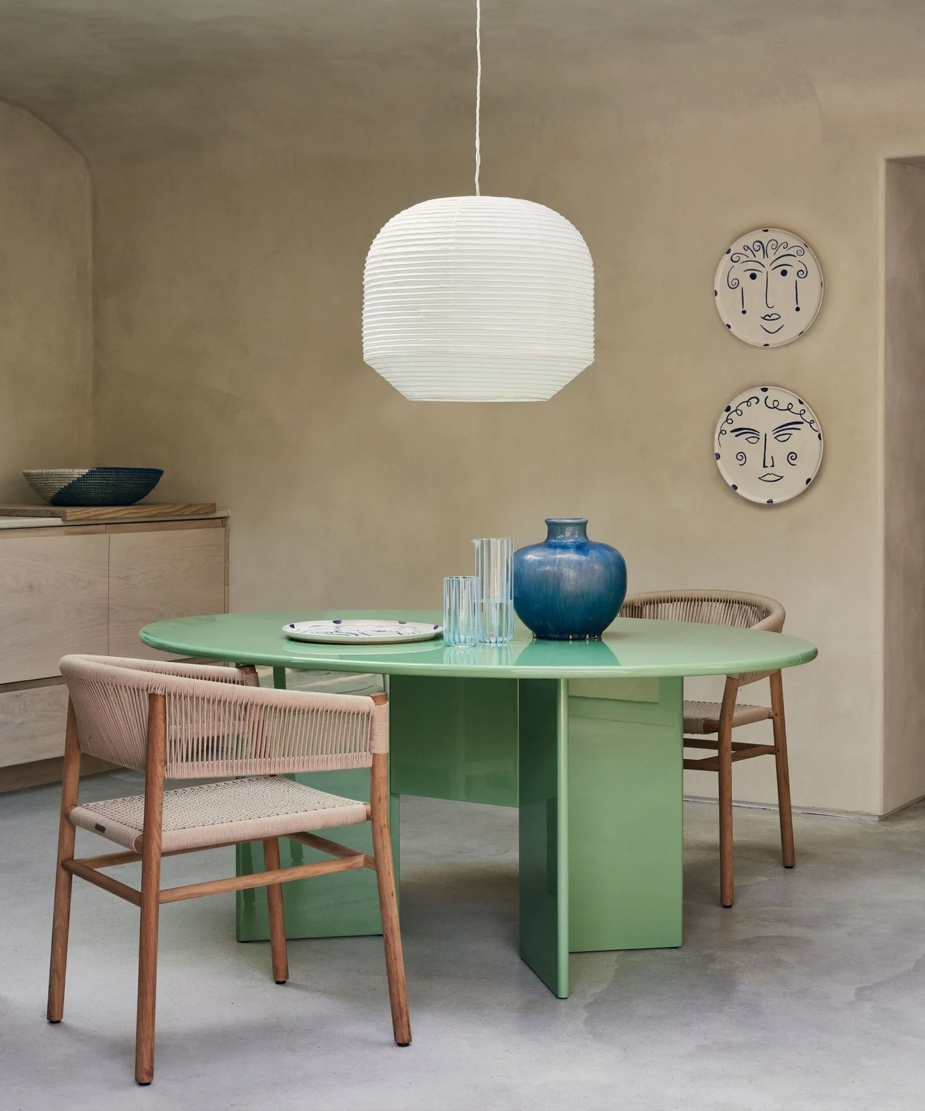 A green round pedestal table sits beside wooden woven-seat chairs, pale cabinets and a white pendant against warm neutral walls