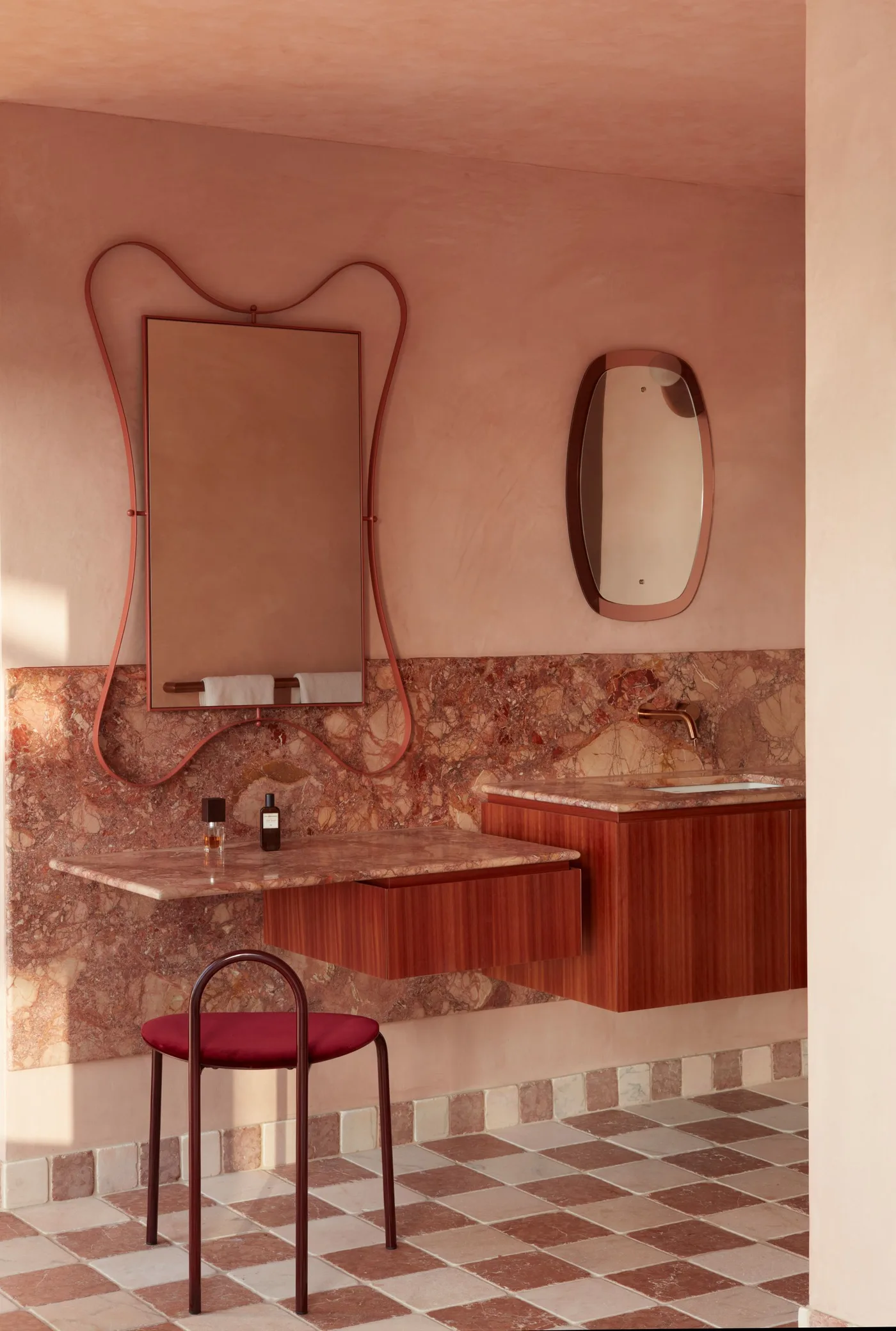 Rose-toned bathroom with patterned counters, floating wood units, a red seat and a pale checker floor