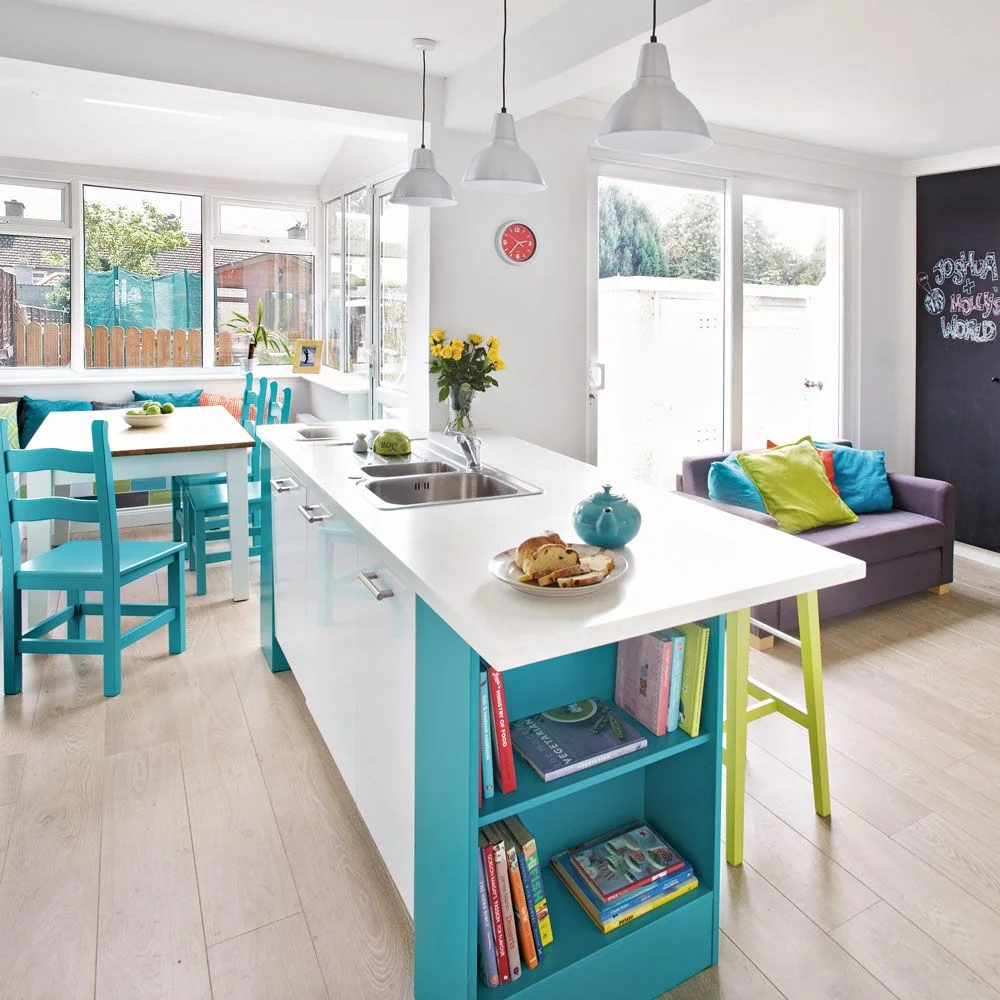 Turquoise dining chairs and a small table sit by broad windows beside a pale kitchen peninsula and turquoise book shelf