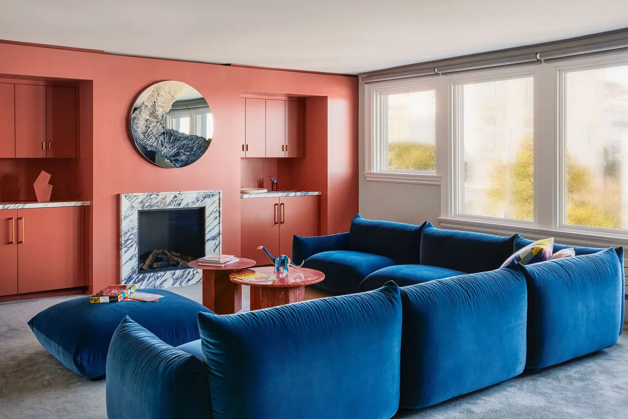A broad blue seating arrangement faces coral-colored built-in storage and a pale fireplace.
