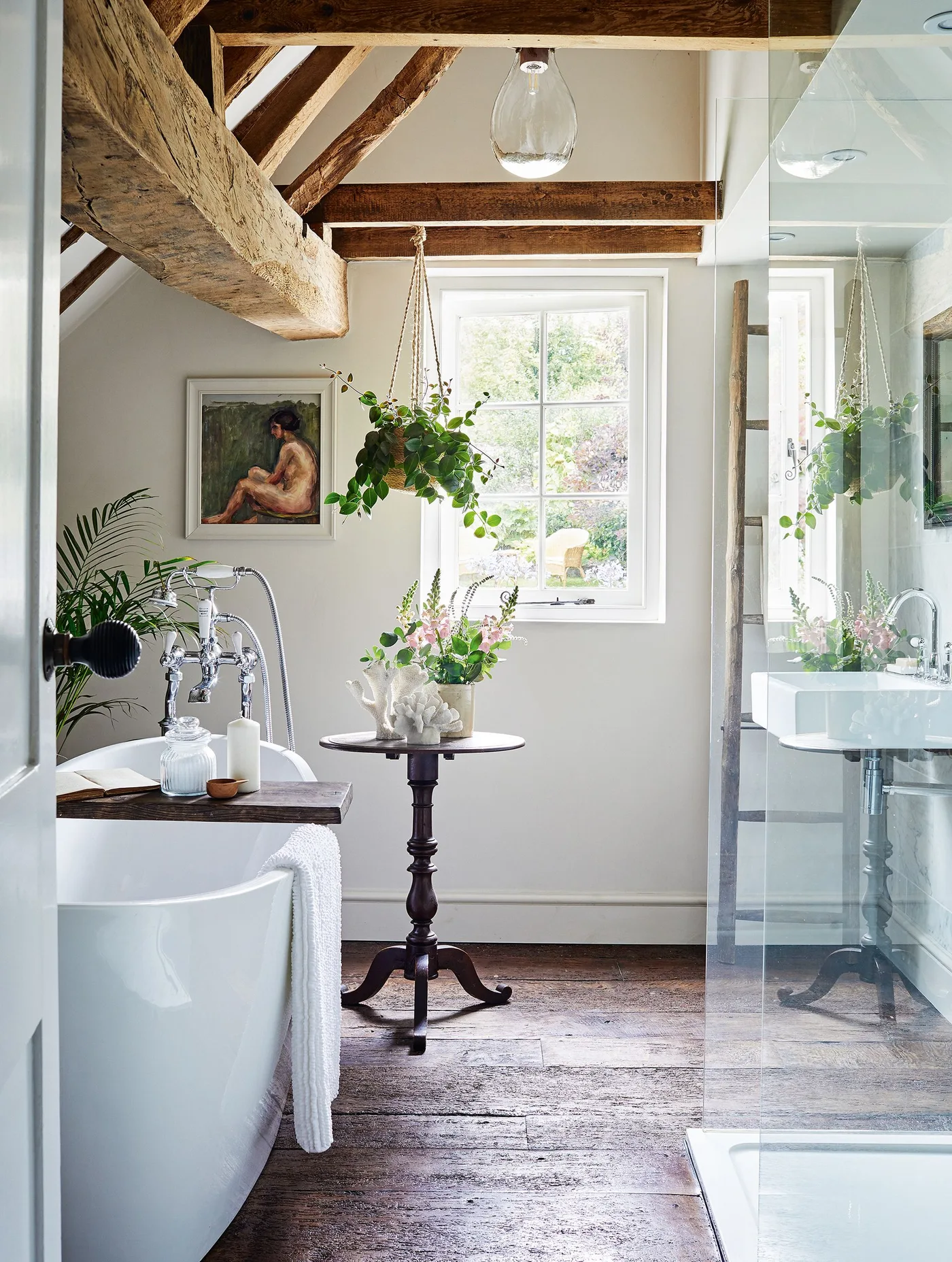 Country bathroom with exposed beams, a white freestanding bath, glass shower screen, hanging plant and wooden floor.