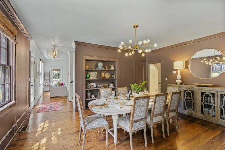 Brown dining-room walls with recessed display shelves beside a doorway, a pale oval table, upholstered chairs, a mirrored-front sideboard, and branching lights.