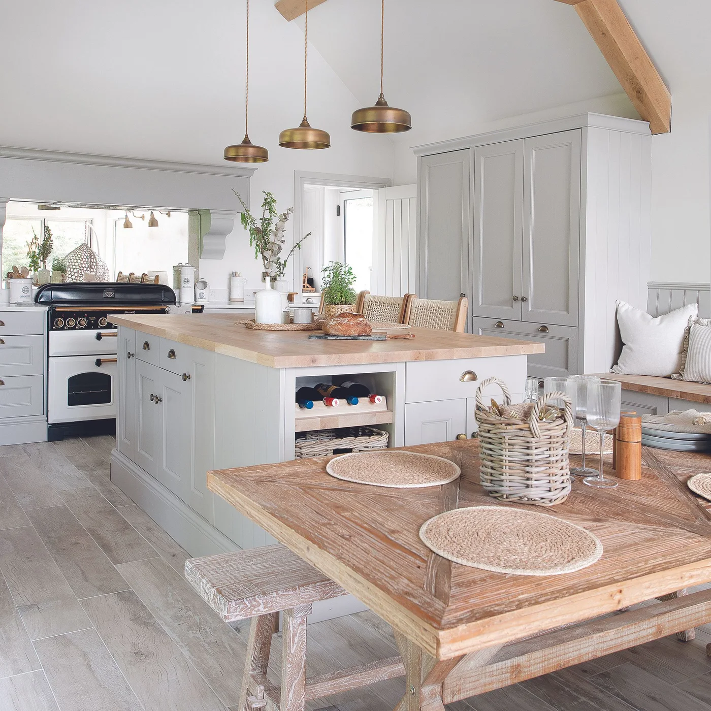 Pale gray cabinets and an island with a wooden top stand beside a wooden dining table beneath a pitched white roof and exposed beam